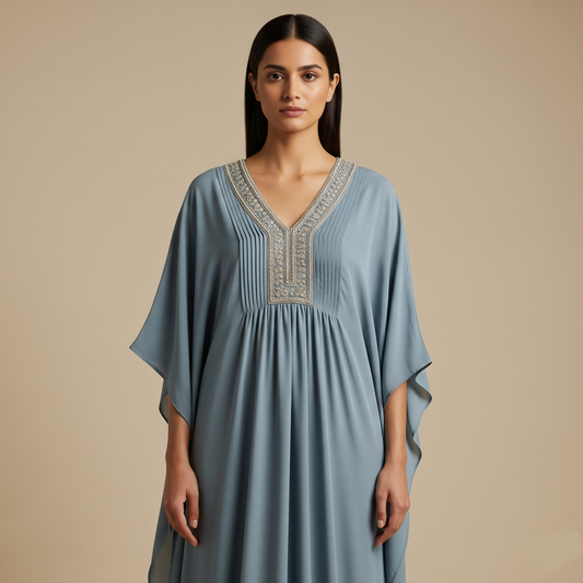A model wears the Alaya Kaftan, a long dusty blue v-neck dress, showcasing its flowing sleeves and intricate silver embroidery.